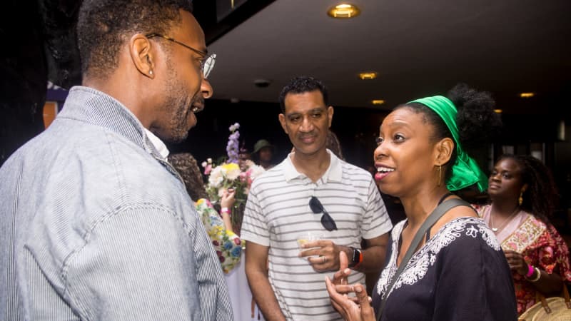 A woman with a green scarf headband speaks to two men among the crowds.