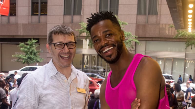 Two men smile together and look at the camera. One wears a button up shirt and a name tag and the other wears a hot pink tank top.