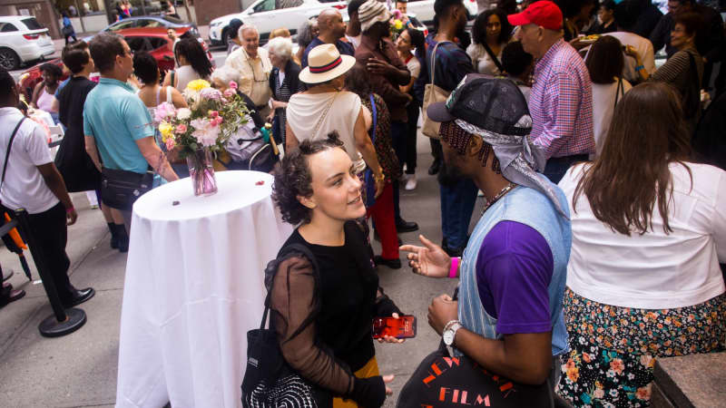 A busy crowd of people talk and stand among high top tables with white table cloths outside the theatre.