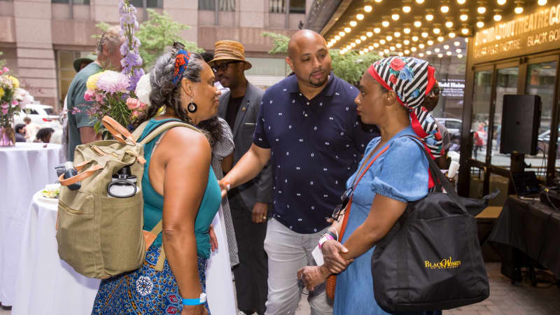Two women wearing scarves in their hair speak to a man outside the theatre underneath the bulbs of the marquee.