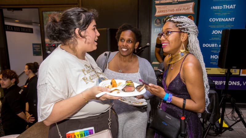 A woman holding a plate of food and with a Playbill sticking out of her bag talks to two other women outside the theatre.