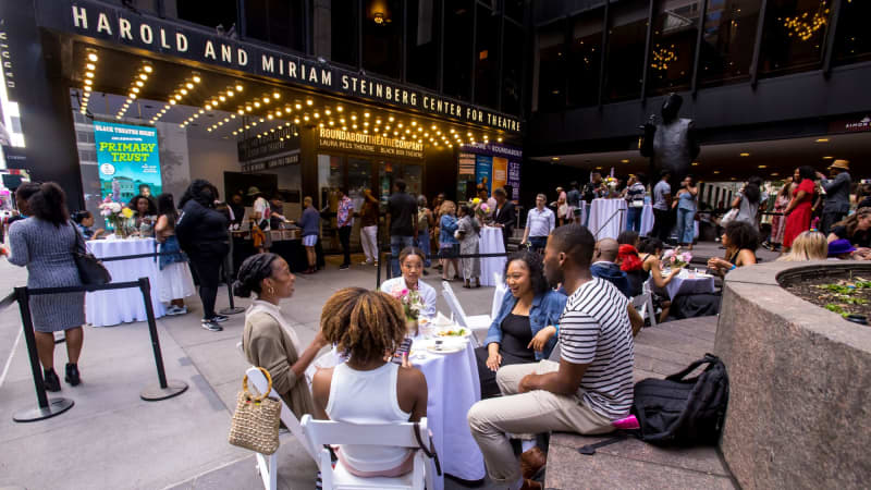 Crowds of people sit at tables and brick benches eating and conversing outside the Harold and Miriam Steinberg Center for Theatre.