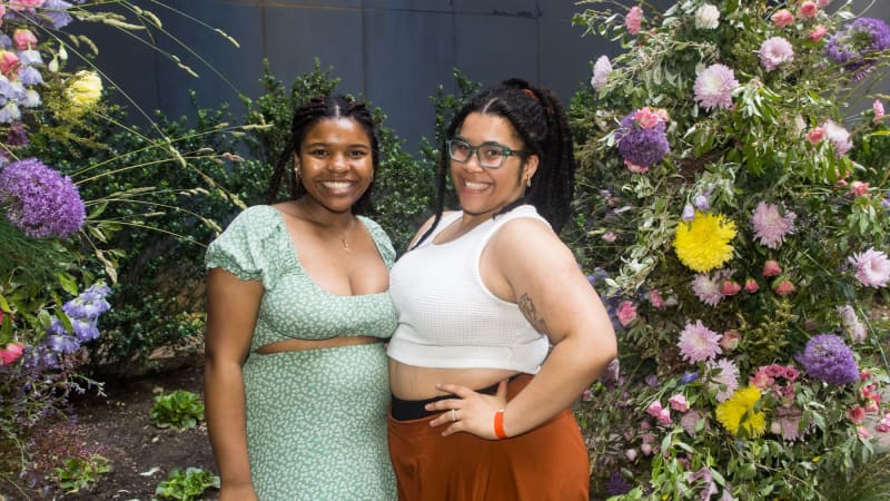 Two people pose together and smile near a large floral arrangement outside.