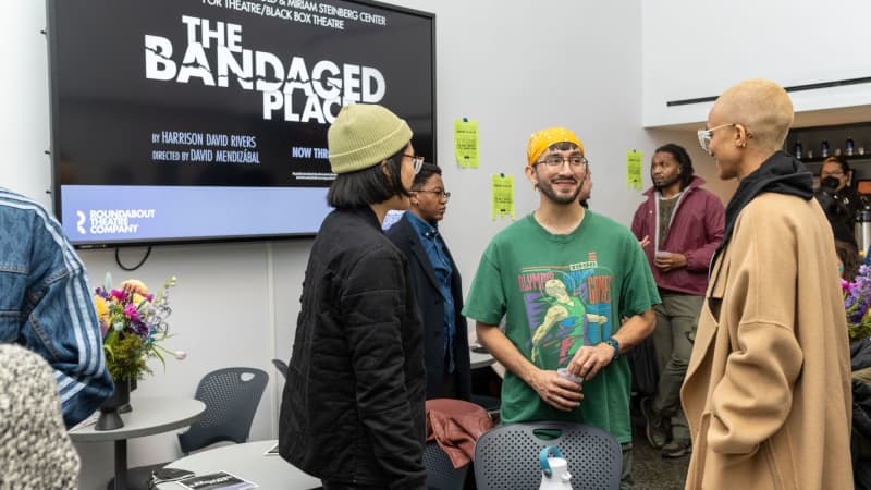 A group of people in a lobby talk in small groups. On the back wall is a large TV with the poster for THE BANDAGED PLACE on it.