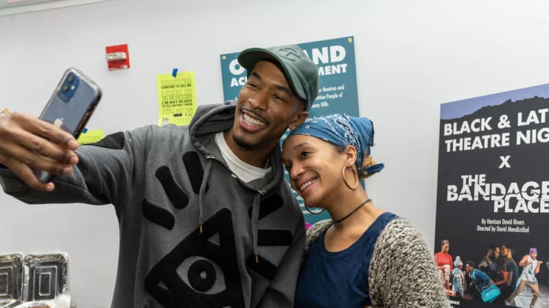 Two people pose for a selfie in front of a sign on an easel that says BLACK AND LATINE THEATRE NIGHT X THE BANDAGED PLACE.