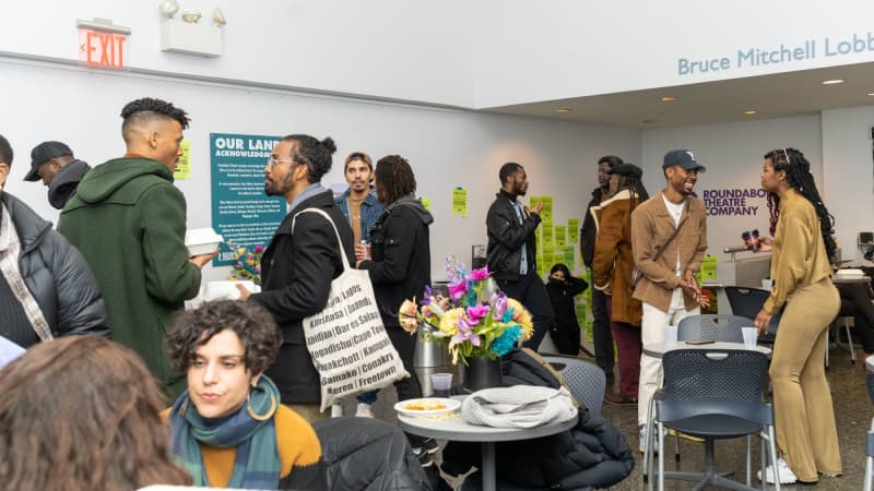 Numerous people stand and sit around a lobby and have conversations with each other. Some hold white takeout containers.