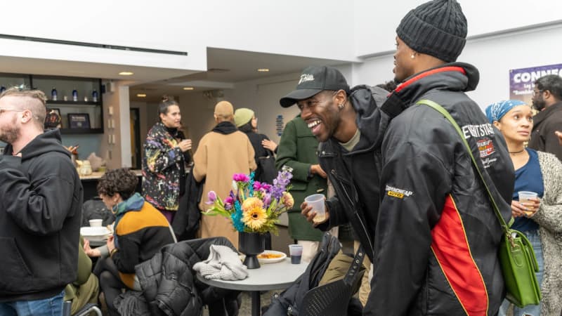 Several people stand and sit in a lobby, holding drinks in plastic cups. One man is caught in the middle of a large laugh.
