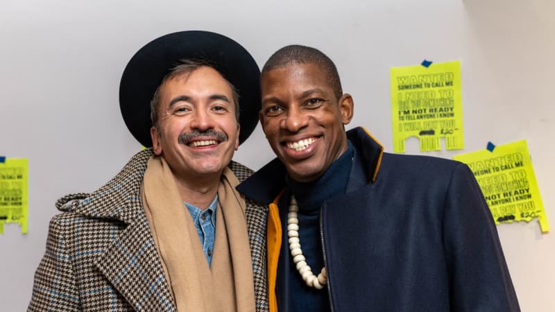 Two people smile for a photo. One has dark grey hair and a mustache, a trench coat, and a black rimmed hat. The other has a shaved head and wears a navy jacket with the collar popped.