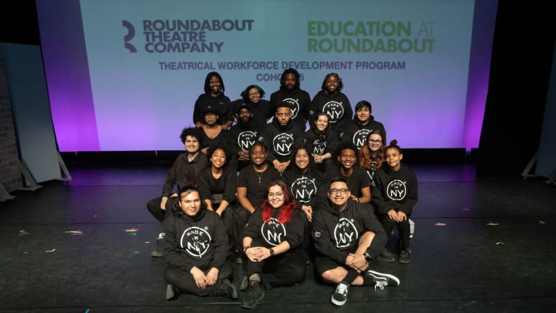 A group of TWDP students wearing black Made in NY hoodies smile together in a clump.