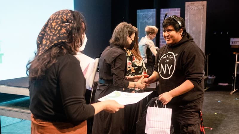 A TWDP student wearing a headset smiles while being handed a certificate.