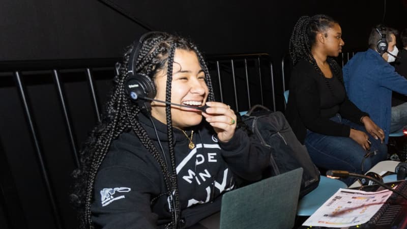 A student wearing a headshot smiles while working behind a lighting board.