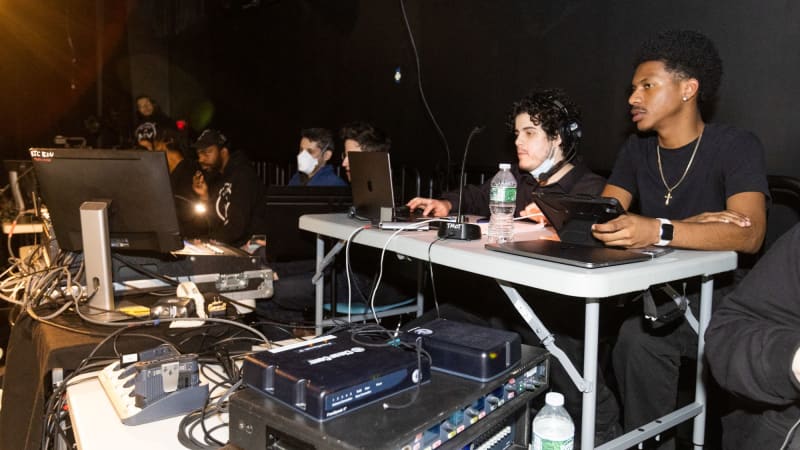 Students in black wear headsets and sit behind a tech table with laptops, sound boards, and microphones.