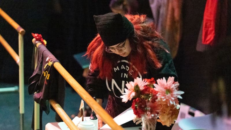 A student in black leans over a prop table to review a script.