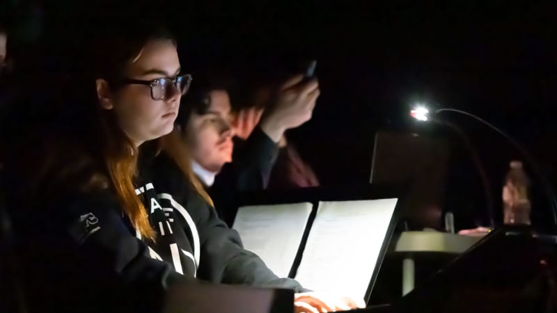 A row of students in the dark review scripts and type on laptops, illuminated by a small light clipped to a music stand.