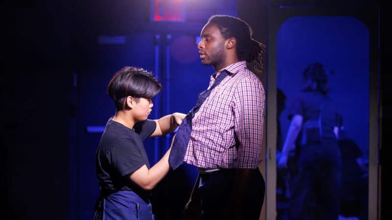 A TWDP fellow dressed in black adjusts the front of a shirt on an actor backstage.