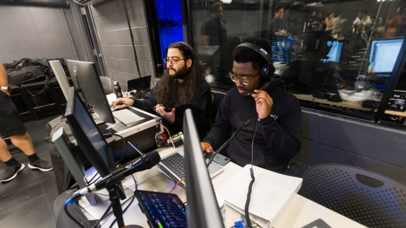 Two TWDP fellows dressed in black wear headsets and sit behind a table full of computers and sound boards.