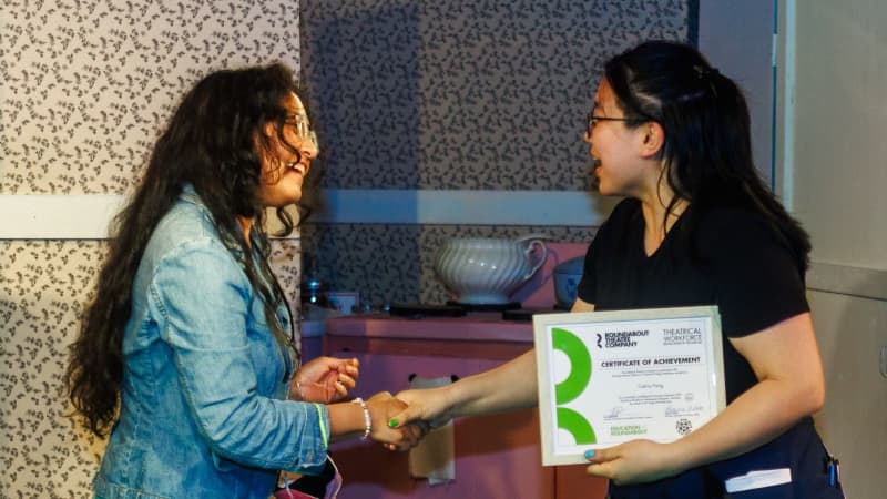 A TWDP student holds a certificate and shakes hands with a Roundabout employee. They both smile.