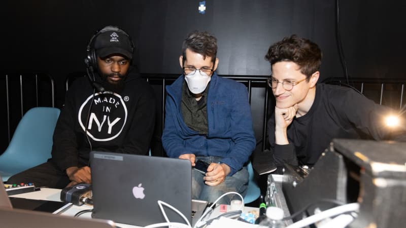 A student in black wearing a headset looks at a laptop with two mentors.