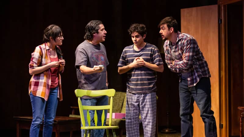 A family of one woman and three men of varying ages gather in a sparse living room with concern.