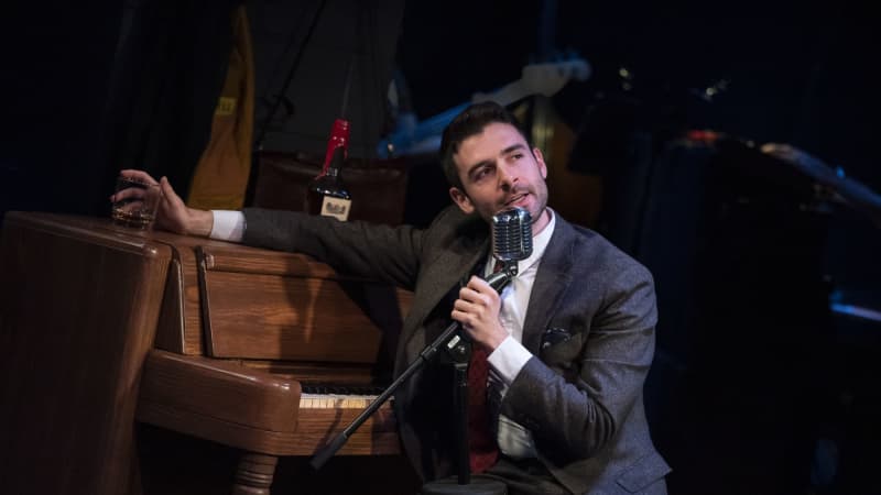 A man in a suit sits at a wooden piano and leans on the top while he pulls a microphone down to his mouth to sing.