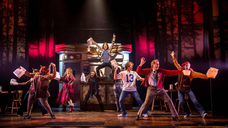 A group of actors joyfully dance with paper bags all around the set of a diner in the middle of the woods with glowing red lights.