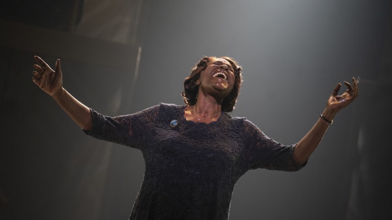 A Black woman in a dark, lacey dress with a blue broach on it and a cross necklace sings passionately with her arms outstretched.