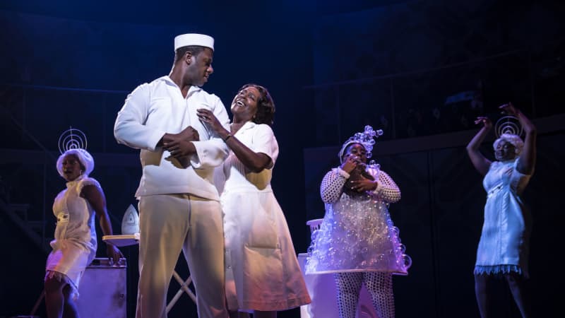 A Black woman in a white maid's dress holds on to and sings to a Black man in a white sailor's uniform. Behind them, the three radio women dance in white whimsical outfits.