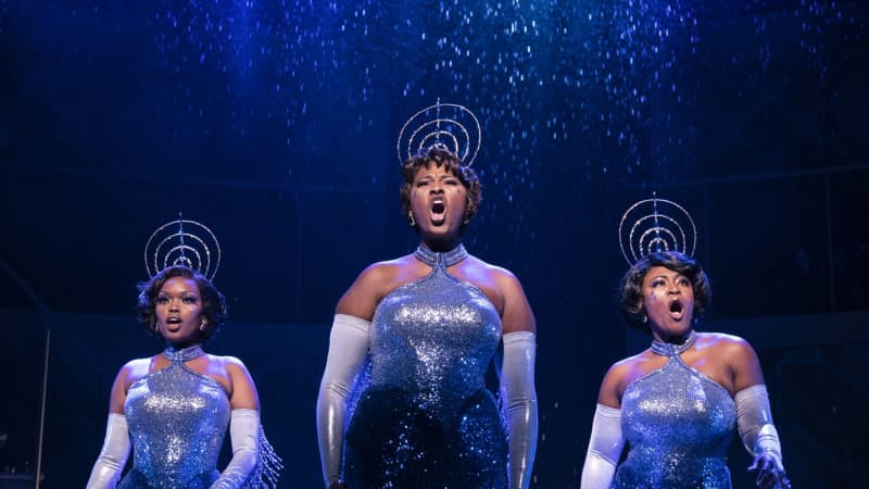 Three Black woman in sleek, sparkly, dark blue dresses with long silver gloves. They wear elaborate, shiny, lagre, silver headbands with layered rings standing on top.