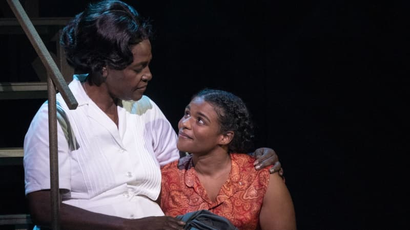 A Black woman in a white maid's dress sits on steps with her arm around a young, smiling Black woman wearing an orange paisley shirt and jeans.