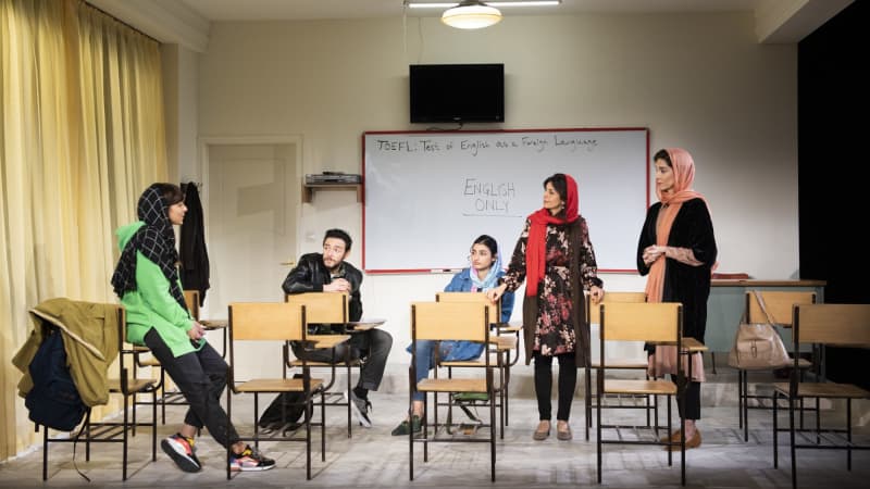 A group of Iranian students and their teacher sit and stand around a classroom. The whiteboard on the wall says, Test of English as a Foreign Language. ENGLISH ONLY.