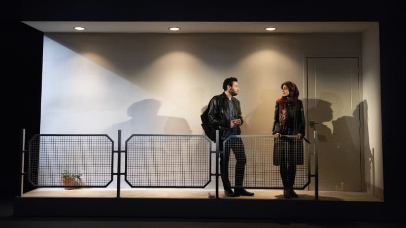 An Iranian man and woman stand on a balcony outside of a gray building with a single door and one small potted plant. They smile at each other.