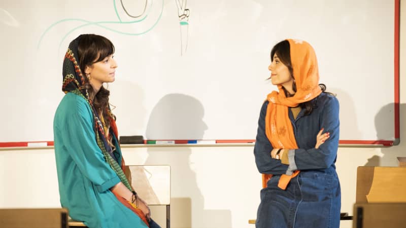 Two Iranian women in stylish hijabs and clothing lean against desks in a classroom. On the whiteboard behind them is an illustration of a hijabi holding up a peace sign with her fingers.