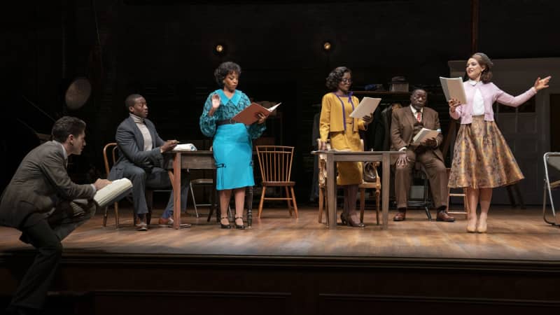 A group of people in 1950s business suits and bright dresses host a rehearsal and read from scripts.