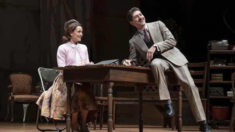 A white woman in a 1950s dress smiles and sits behind a desk, which a white man in a business suit smiles and half-sits on top of.