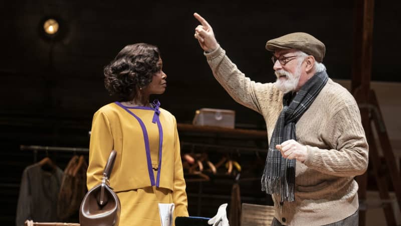 An old white man with a white beard raises a pointed finger and speaks to a Black woman in a yellow suit dress.