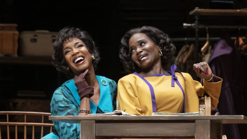 A Black woman in a bright turquoise dress and a Black woman in a bright yellow dress sit next to each other and sing gleefully.