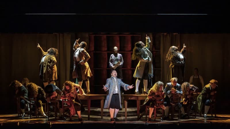 Actors performing "Molasses to Rum." Edward Rutledge stands center with arms outstretched. Jefferson stands on a barrel behind him, in the shadow of actors standing on two tables with arms pointed up.