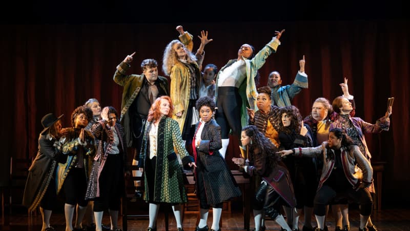 Company of 1776 on stage. Actors all looking and pointing in different directions, all with anger towards John Adams, who is in the center. Half the company is standing on tables and chairs. 