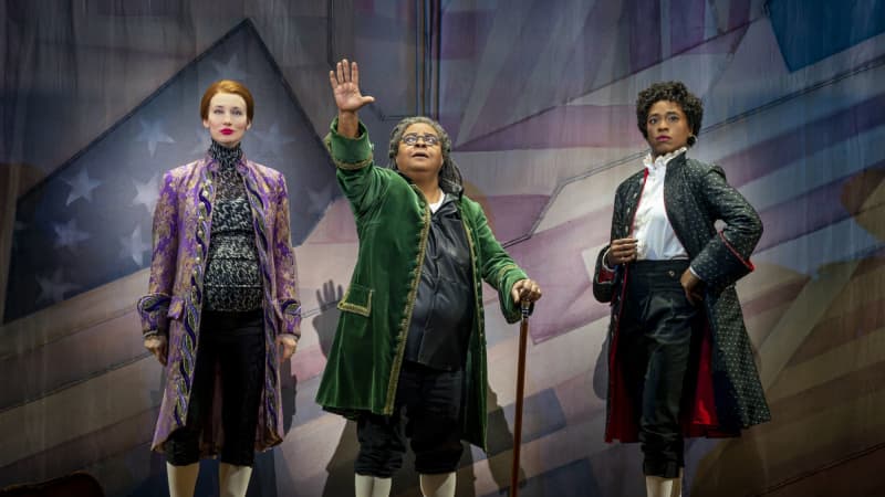 Thomas Jefferson, Benjamin Franklin, and John Adams stand in front of a curtain patched with stripes from the American flag and stare into the distance. Franklin extends an arm outwards.