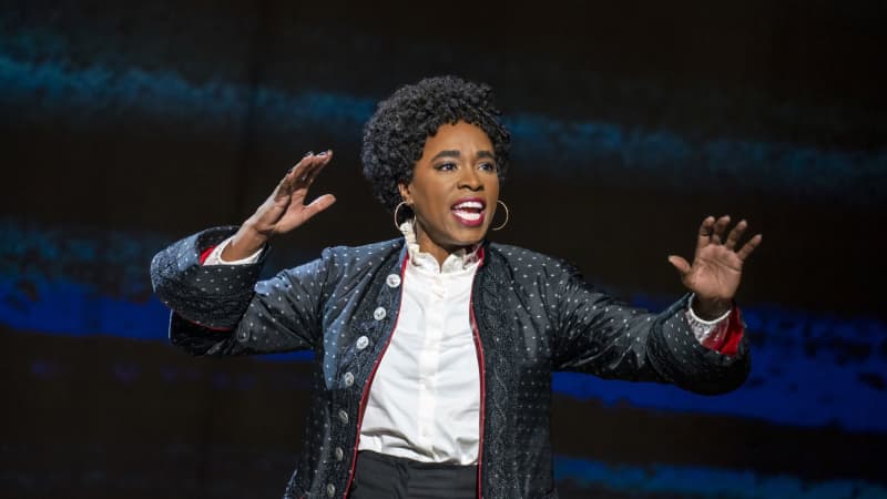 Kristolyn Lloyd as John Adams singing passionately. She has dark skin, short black hair, and wears a frilly white button down underneath a fancy black coat with a red lining.