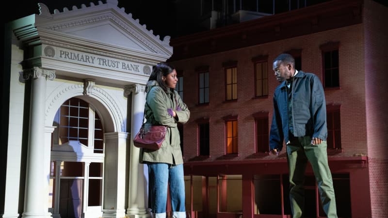 A woman and man stand across from each other in front of scaled-down buiilding fronts, one of which is a bank. The woman bundles up with her jacket.