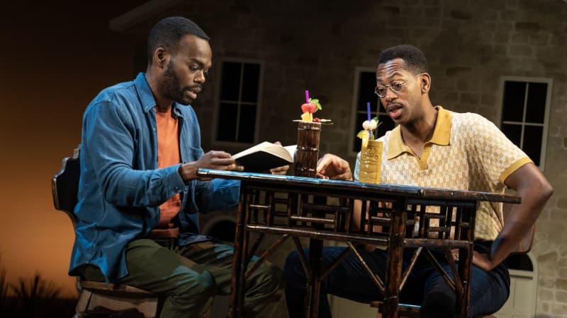 Two men sit at a wooden table with colorful, tropical drinks on top. One of them reads from a book.