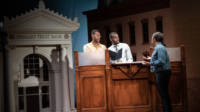 Two men stand behind a bank counter and listen to a woman who is their customer. Behind them is a scaled-down front of a bank named PRIAMRY TRUST BANK.