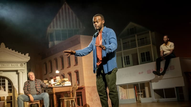 A man with dark skin stands center stage talking to the audience. Behind him on either side in the distance sits an older white man on the left and a Black man on the right, sitting on a rooftop.