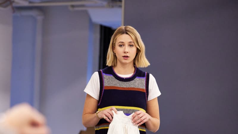 A woman with short blonde hair above her shouldres stands in front of a table with a laundry basket on top. She holds a white baby nightgown. 