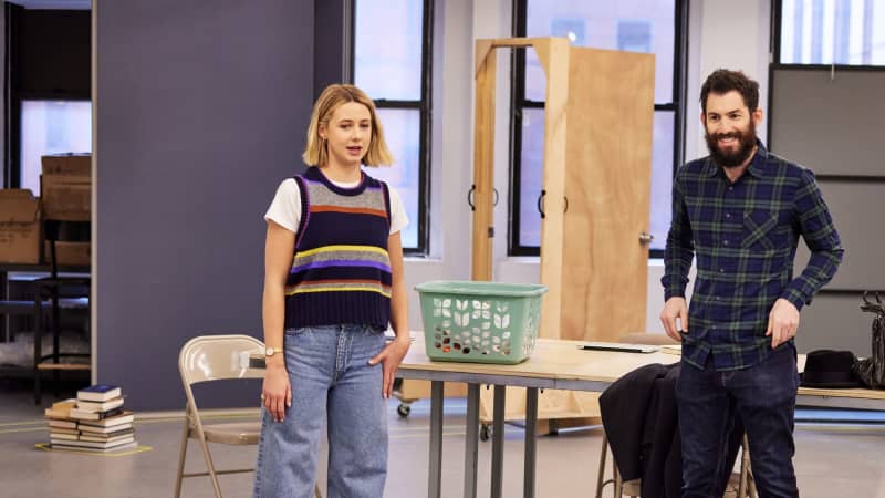 A woman and a man stand in front of a long table with a laundry basket on top. They're looking past the camera at the director's table. Stacks of books are scattered across the floor.