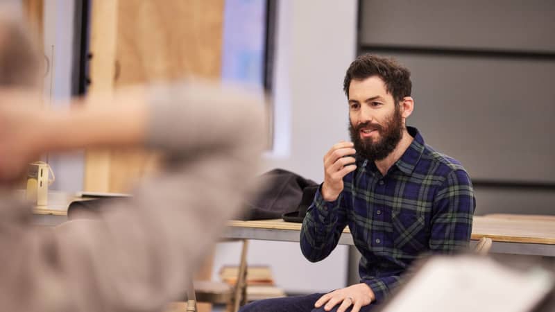 A man sits in front of a long table in the rehearsal studio and reaches a hand up towards his face. He has dark brown hair, a full beard, and wears a green flannel shirt and jeans.