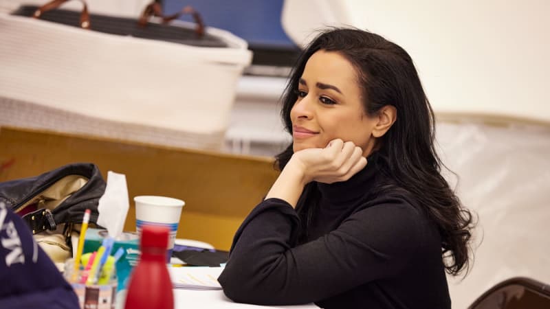 A smiling woman with mid-length black hair sits at a messy table in the rehearsal studio. She wears a black turtleneck and props an elbow on her script, resting her head on her hand.