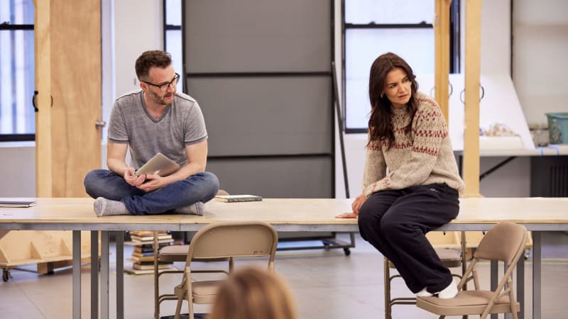 A man and a woman sit on top of a long table on oppostite sides. He is cross-legged and she sits with her legs hanging off the table, feet resting on the chair below her.