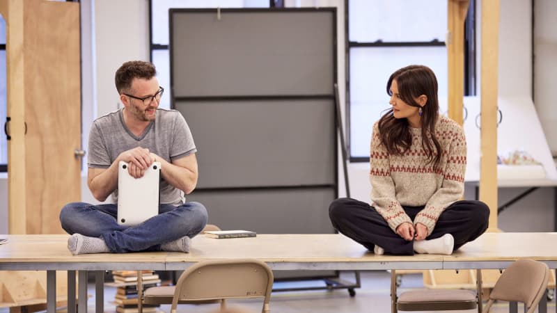 A man and a woman sit cross-legged on opposite ends of a long table. The man holds a closed laptop in his lap and smiles at the woman.